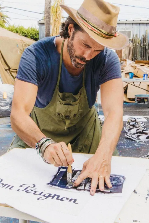 Greg Lauren painting a canvas sign in his studio, wearing a hat and apron